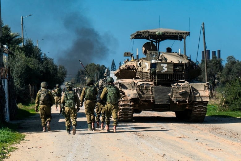 Helmeted soldiers are seen from behind walking beside a tank-like vehicle. In the skyline ahead, black smoke is shown.