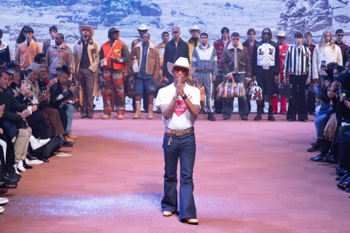 A man in a wide-brimmed white hat, shades and wide-flared jeans on a fashion catwalk